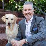 Man in a grey suit sitting next to a golden labrador
