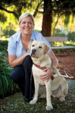 Susan Armstrong in a park kneeling next to a yellow lab wearing a walking harness.